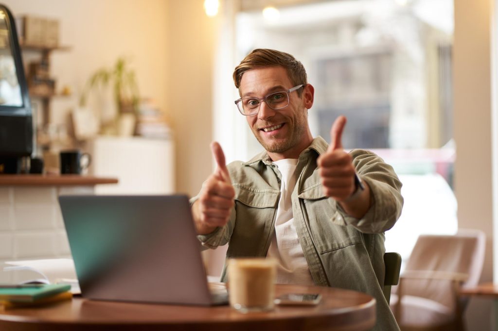 Joyful smiling guy in glasses man shows thumbs up sits in cafe with laptop recommends website