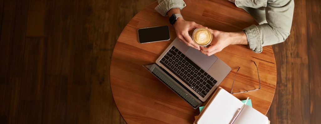 Top view male hands holding cup of coffee man sitting at round table in a cafe working on laptop had his smartphone and notebook drinking cappuccino