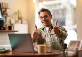 Joyful smiling guy in glasses man shows thumbs up sits in cafe with laptop recommends website